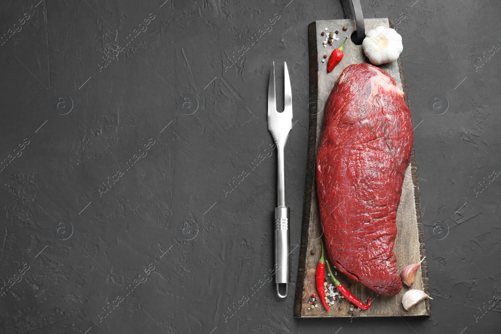 Photo of Piece of raw beef meat, garlic, chili, spices and fork on grey textured table, flat lay. Space for text