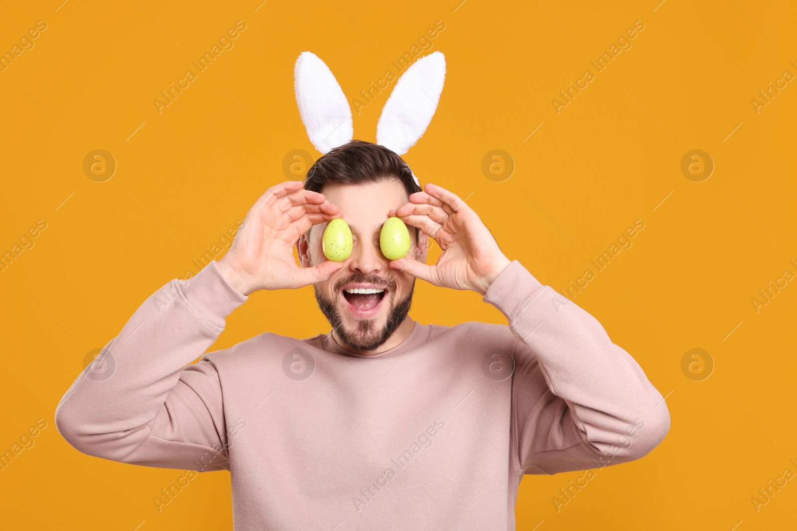 Photo of Happy man in bunny ears headband holding painted Easter eggs near his eyes on orange background