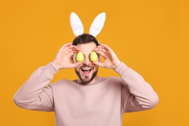 Happy man in bunny ears headband holding painted Easter eggs near his eyes on orange background