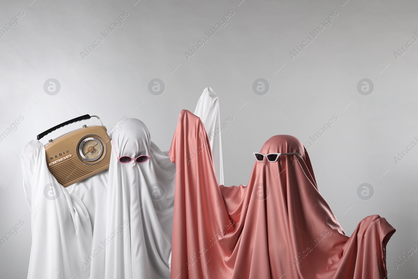 Photo of Glamorous ghosts. Women in color sheets and sunglasses with retro radio receiver on light grey background