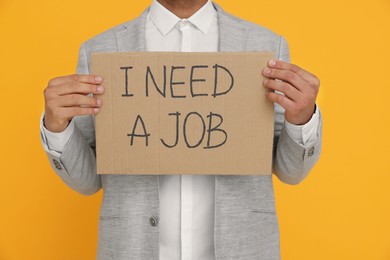 Photo of Young unemployed man holding sign with phrase I Need A job on yellow background, closeup