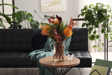 Vase with bouquet of beautiful leucospermum flowers and air freshener on side table near black sofa in room