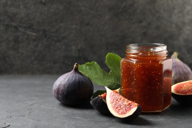 Photo of Glass jar with tasty sweet jam, green leaf and fresh figs on grey textured table. Space for text
