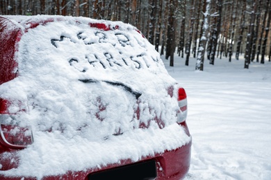 Photo of Phrase "Merry Christmas" written on snow covered car in winter forest. Space for text