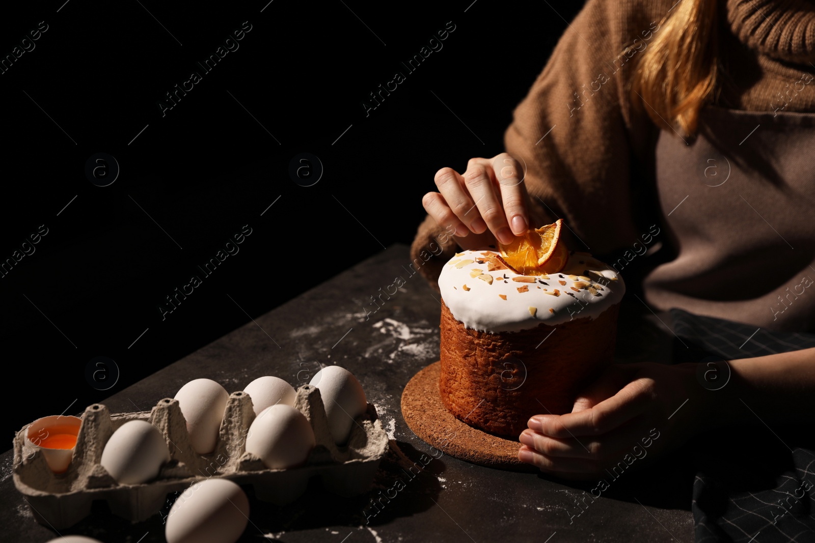 Photo of Young woman decorating traditional Easter cake at table against black background, closeup. Space for text