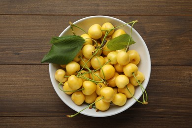 Bowl with ripe yellow cherries and green leaves on wooden table, top view