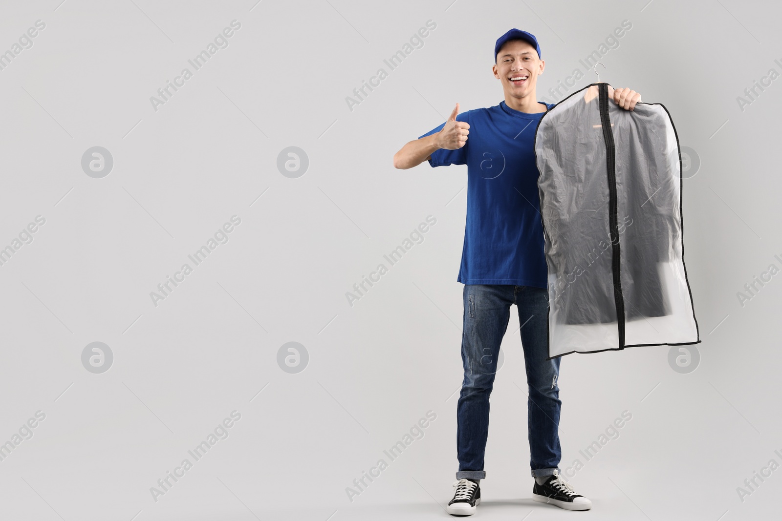 Photo of Dry-cleaning delivery. Happy courier holding garment cover with clothes and showing thumbs up on light grey background, space for text