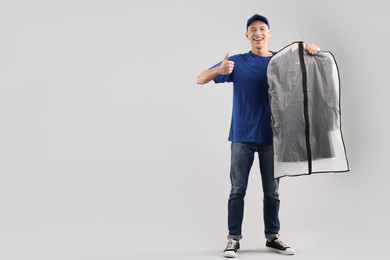 Dry-cleaning delivery. Happy courier holding garment cover with clothes and showing thumbs up on light grey background, space for text