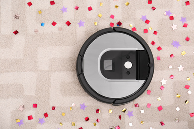 Photo of Modern robotic vacuum cleaner removing confetti from carpet, top view