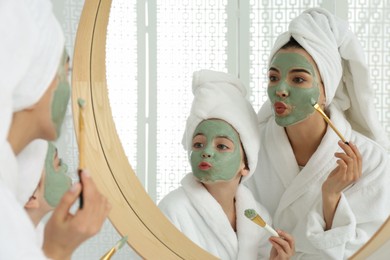 Photo of Young mother and her daughter with facial masks having fun near mirror in bathroom