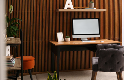 Comfortable workplace with computer near wooden wall in stylish room interior. Home office design