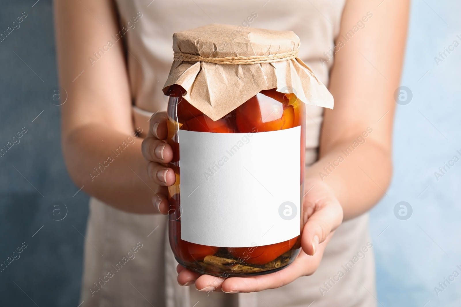 Photo of Woman holding glass jar of pickled tomatoes with blank sticker on blue background, closeup view