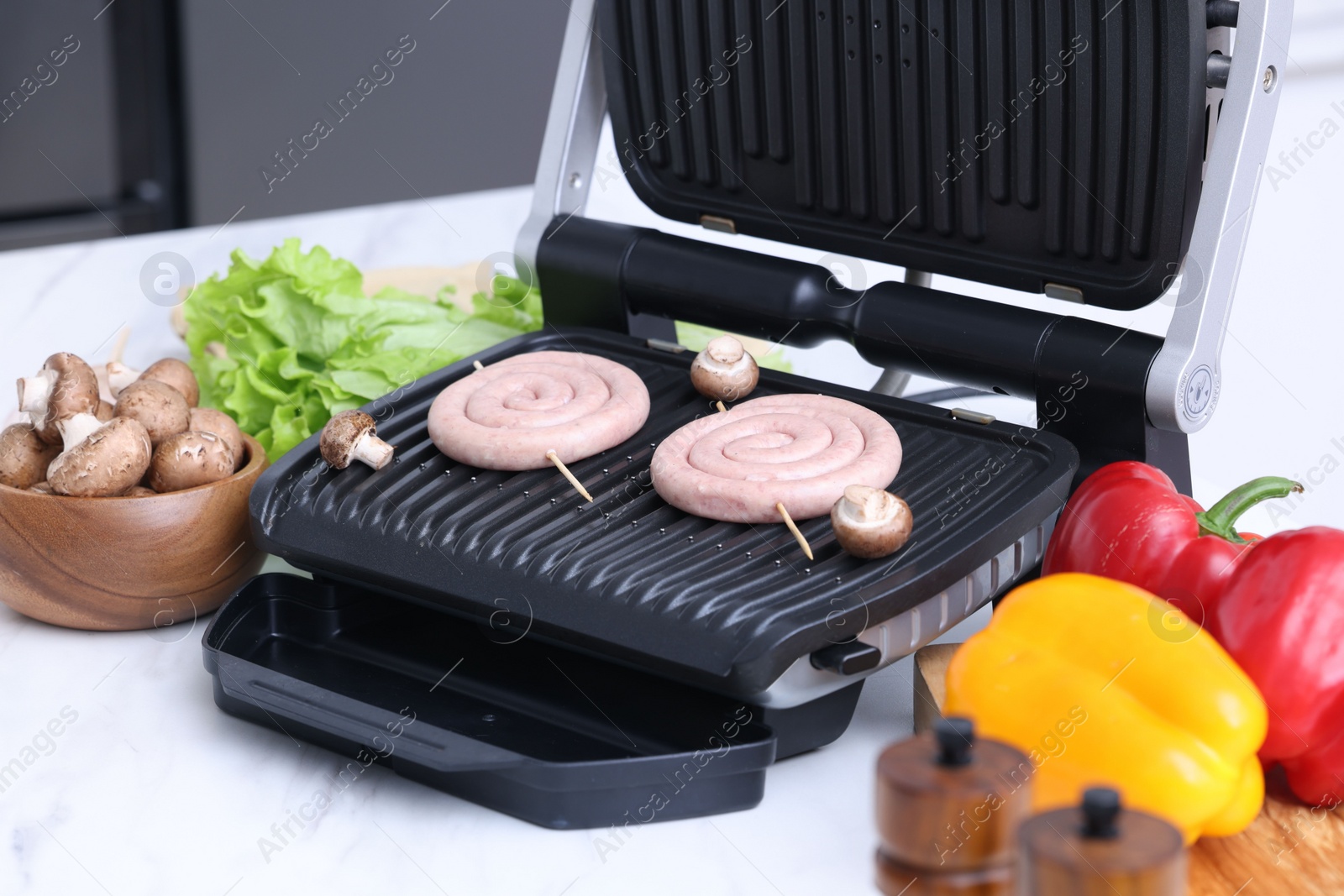 Photo of Electric grill with homemade sausages, mushrooms, and vegetables on white table indoors