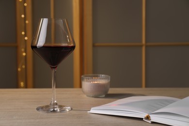 Glass of red wine, open book and candle on wooden table indoors, space for text