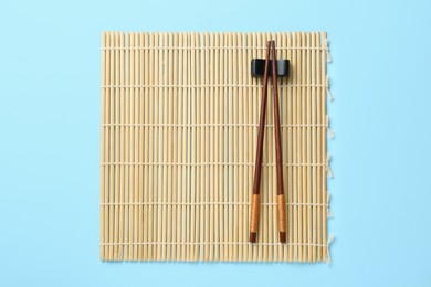 Photo of Bamboo mat with pair of wooden chopsticks and rest on light blue background, top view