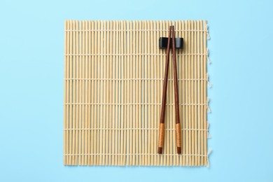 Bamboo mat with pair of wooden chopsticks and rest on light blue background, top view