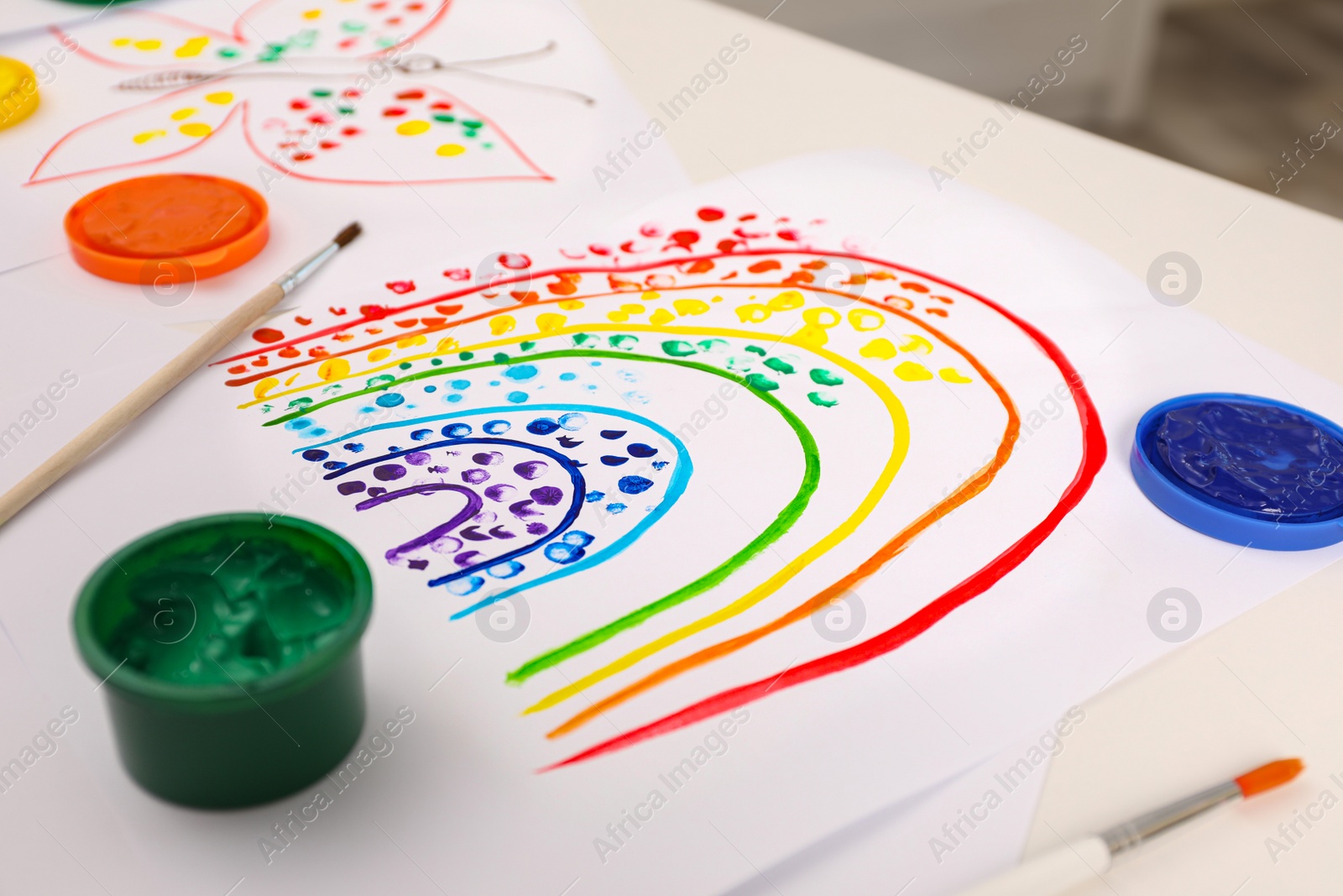 Photo of Cute children's drawing, brushes and set of paints on white table, closeup