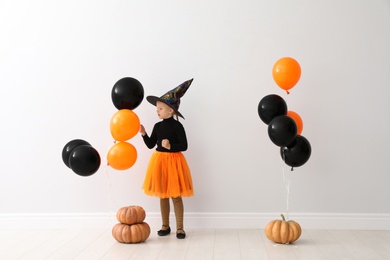 Photo of Cute little girl with balloons and pumpkins wearing Halloween costume near light wall