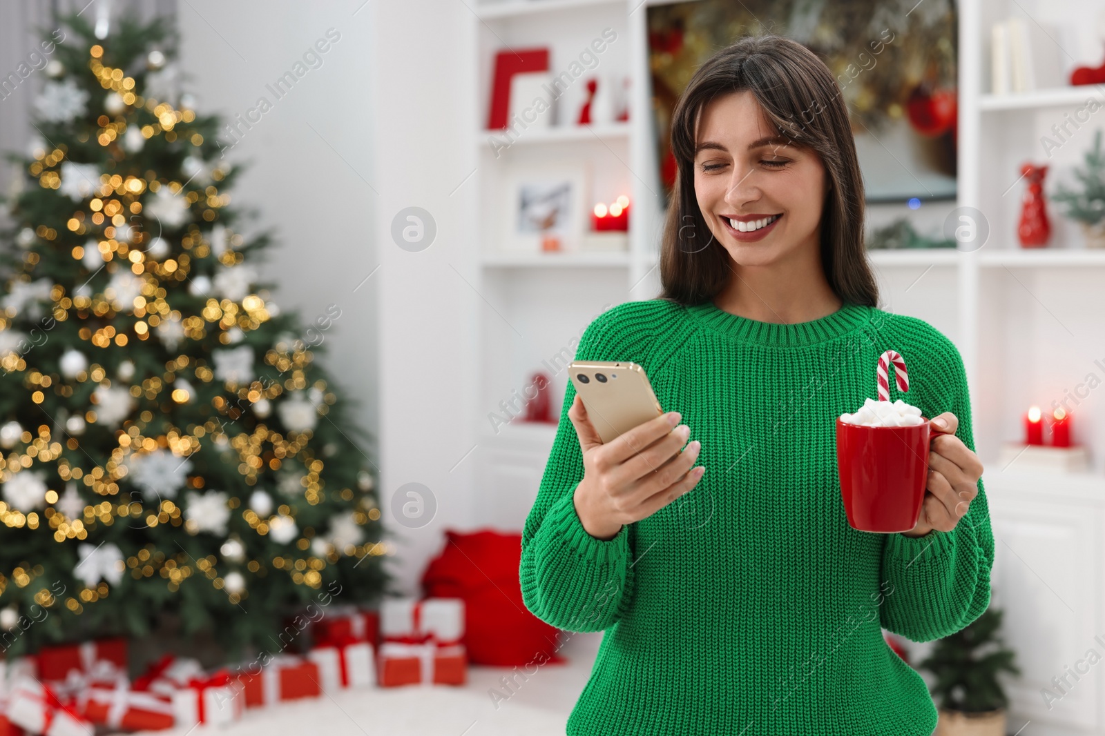 Photo of Smiling woman with cup of Christmas cocoa and smartphone at home. Space for text