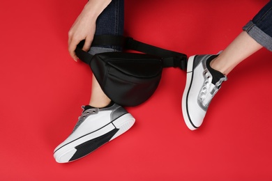 Woman in stylish shoes with bum bag sitting on color background