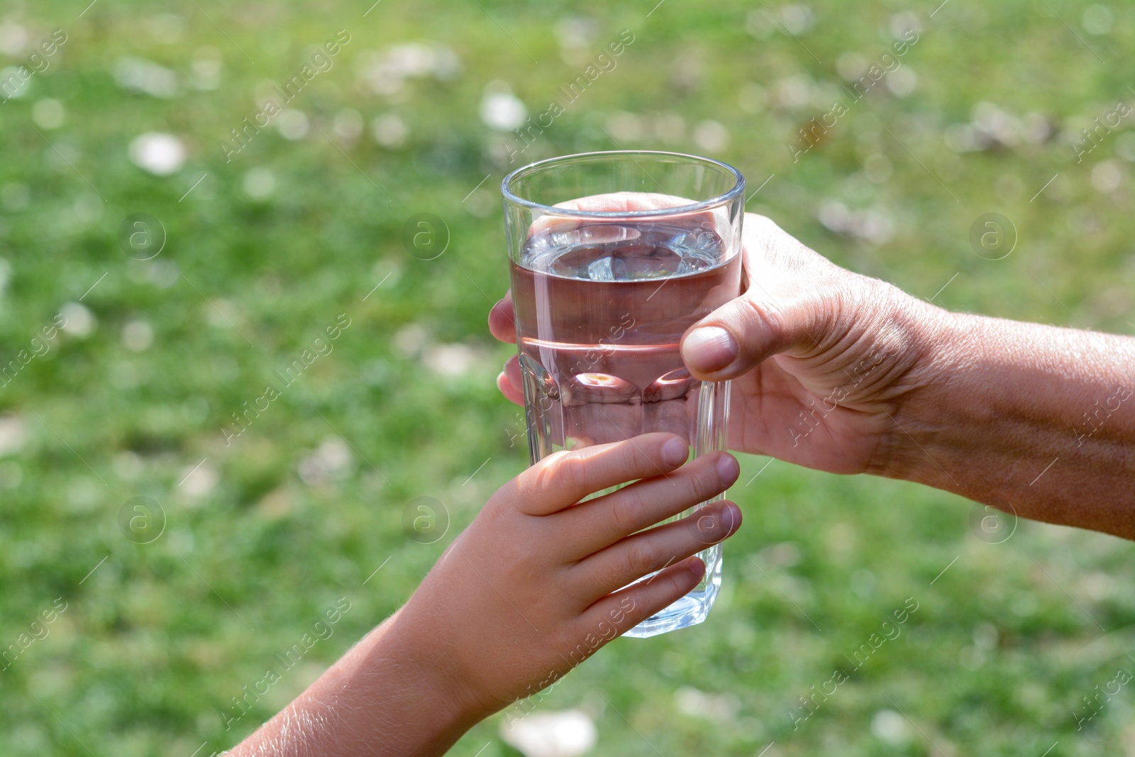 Photo of Child giving glass of water to elderly woman outdoors, closeup