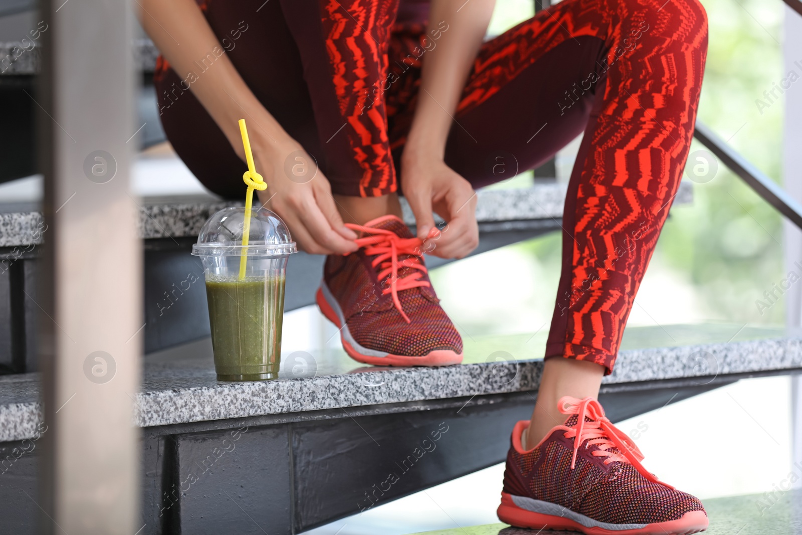 Photo of Young woman in sportswear with plastic cup of healthy smoothie on stairs indoors