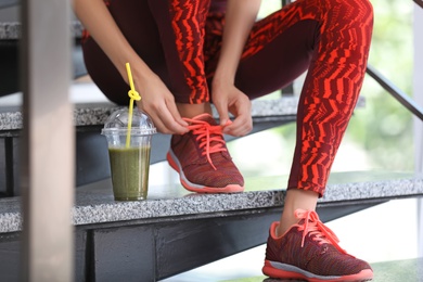 Photo of Young woman in sportswear with plastic cup of healthy smoothie on stairs indoors