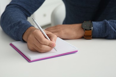 Man writing in notebook at white table indoors, closeup