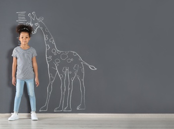Photo of African-American child measuring height near chalk giraffe drawing on grey wall. Space for text