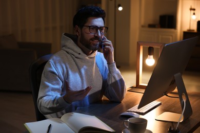 Home workplace. Man talking on smartphone while working with computer at wooden desk at night
