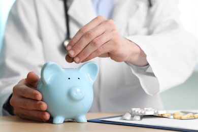 Photo of Doctor putting coin into piggy bank at wooden table, closeup