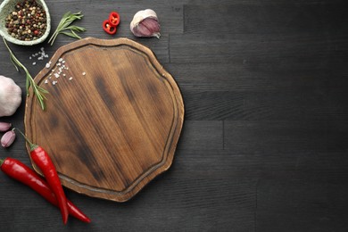 Photo of Cutting board, spices, garlic, rosemary and chili peppers on black wooden table, flat lay. Space for text