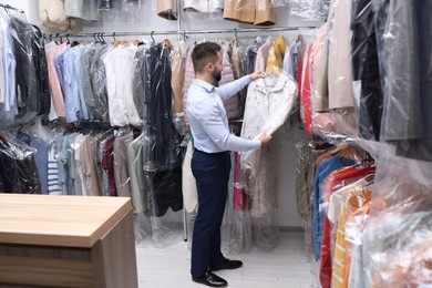 Photo of Dry-cleaning service. Worker holding hanger with jacket in plastic bag near other clothes indoors