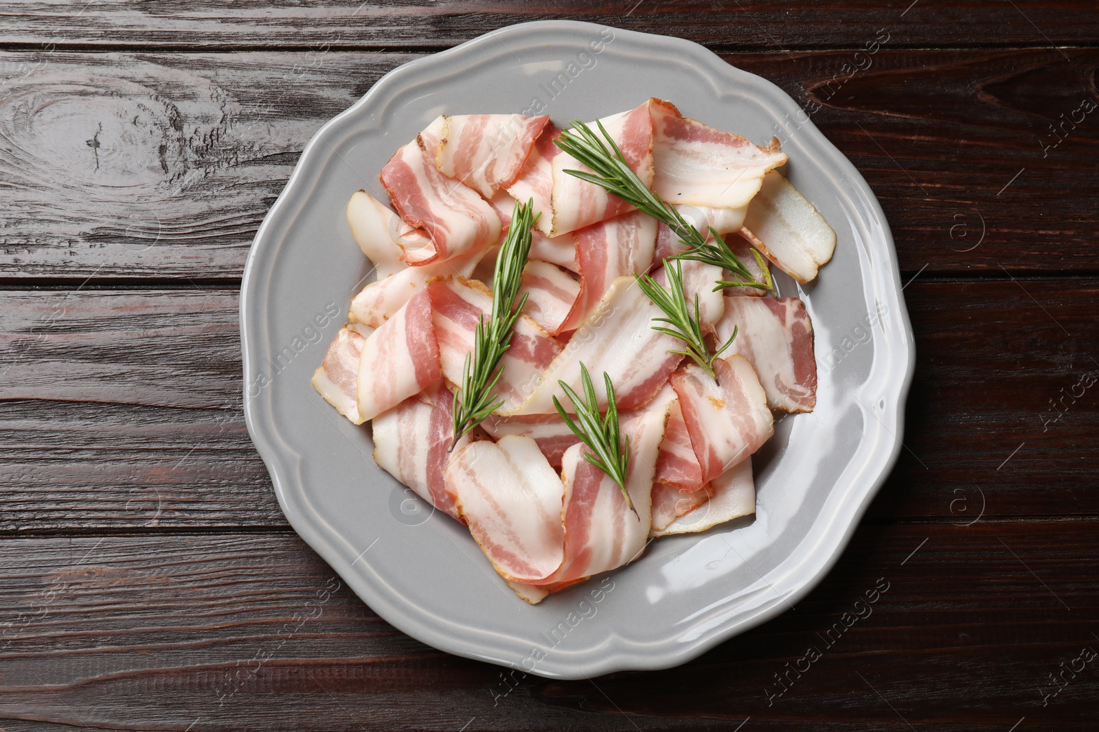 Photo of Slices of raw bacon and rosemary on dark wooden table, top view