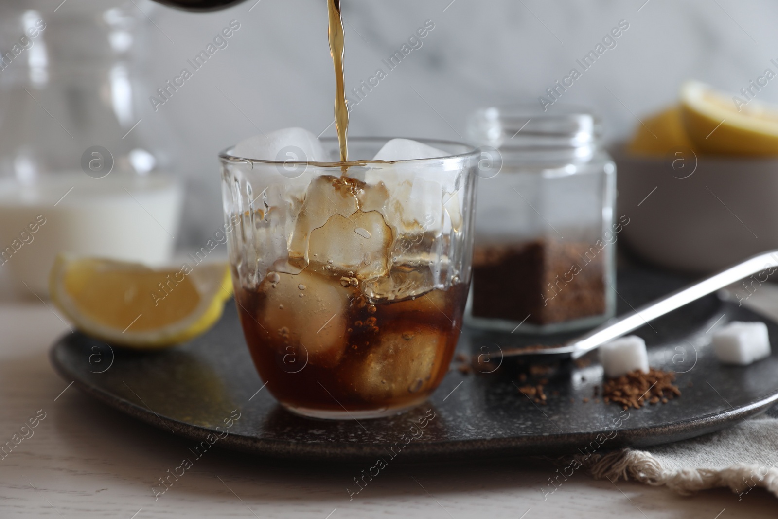 Photo of Pouring coffee into glass with ice cubes at white wooden table, closeup