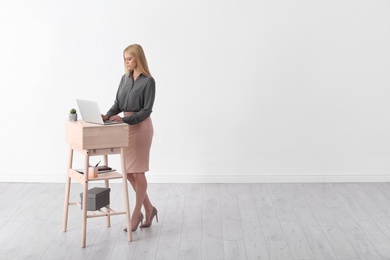 Young woman using laptop at stand up workplace against white wall. Space for text