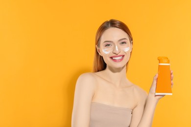Photo of Beautiful young woman with sun protection cream on her face holding sunscreen against orange background, space for text
