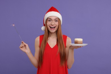 Photo of Emotional young woman in Santa hat with piece of cake and burning sparkler on purple background