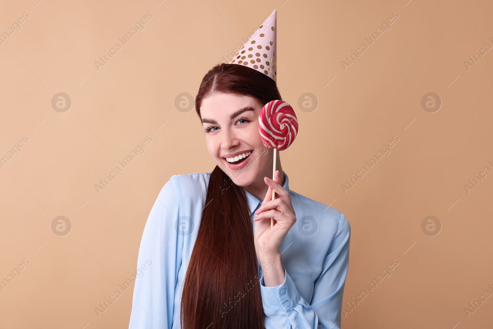 Photo of Happy woman in party hat with lollipop on beige background