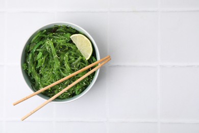 Photo of Tasty seaweed salad in bowl served on white tiled table, top view. Space for text