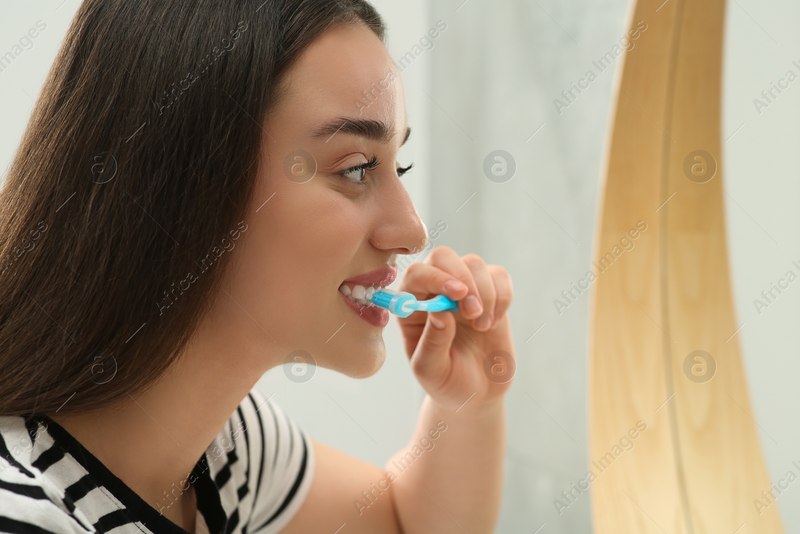 Photo of Young woman brushing her teeth with plastic toothbrush near mirror in bathroom