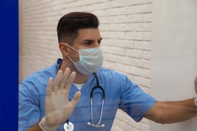 Photo of Sad doctor near window, view from outdoors. Stress of health care workers during coronavirus pandemic