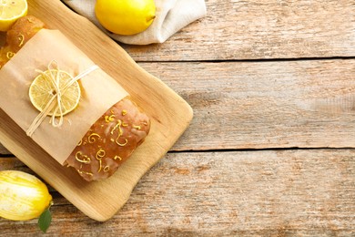 Photo of Wrapped tasty lemon cake with glaze and citrus fruits on wooden table, flat lay. Space for text