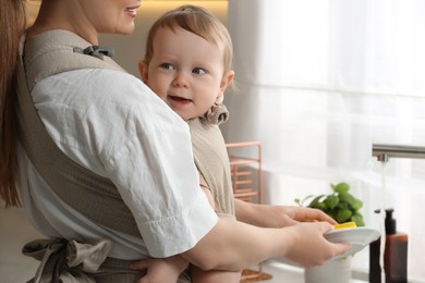 Photo of Mother holding her child in sling (baby carrier) while washing plates at home