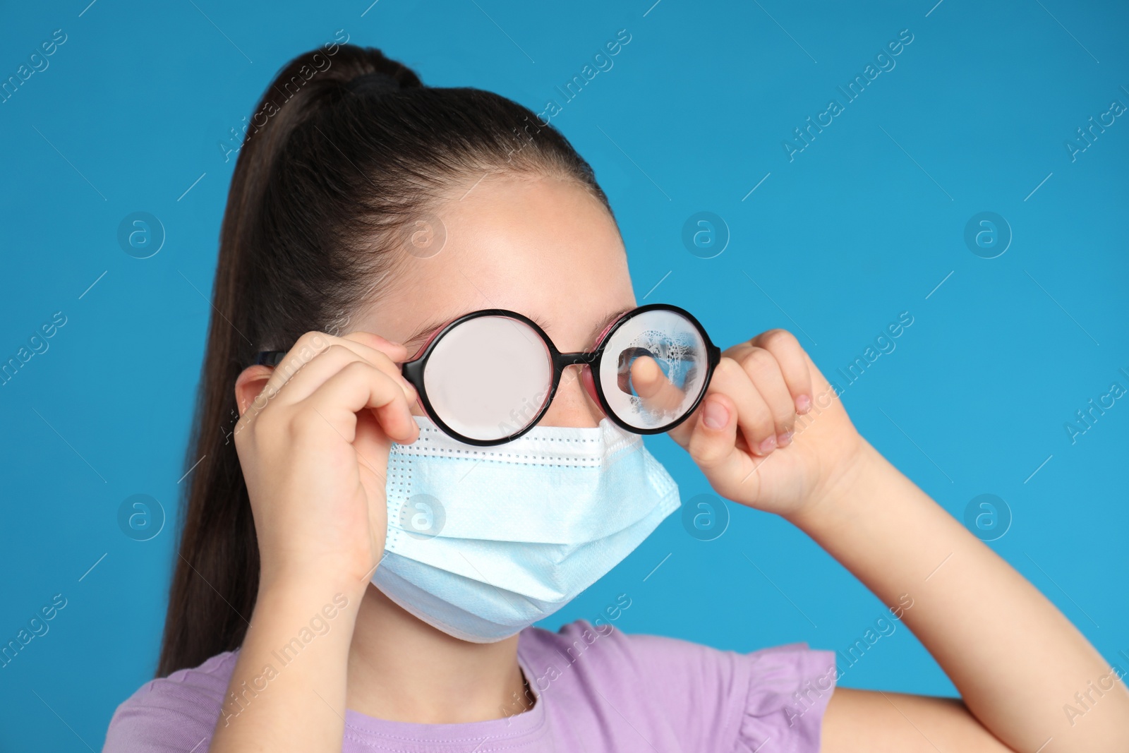 Photo of Little girl wiping foggy glasses caused by wearing medical face mask on blue background. Protective measure during coronavirus pandemic