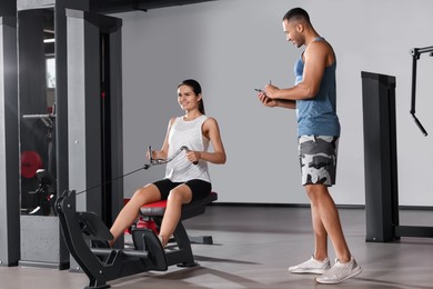 Happy trainer writing down plan of workouts while woman doing exercise in modern gym