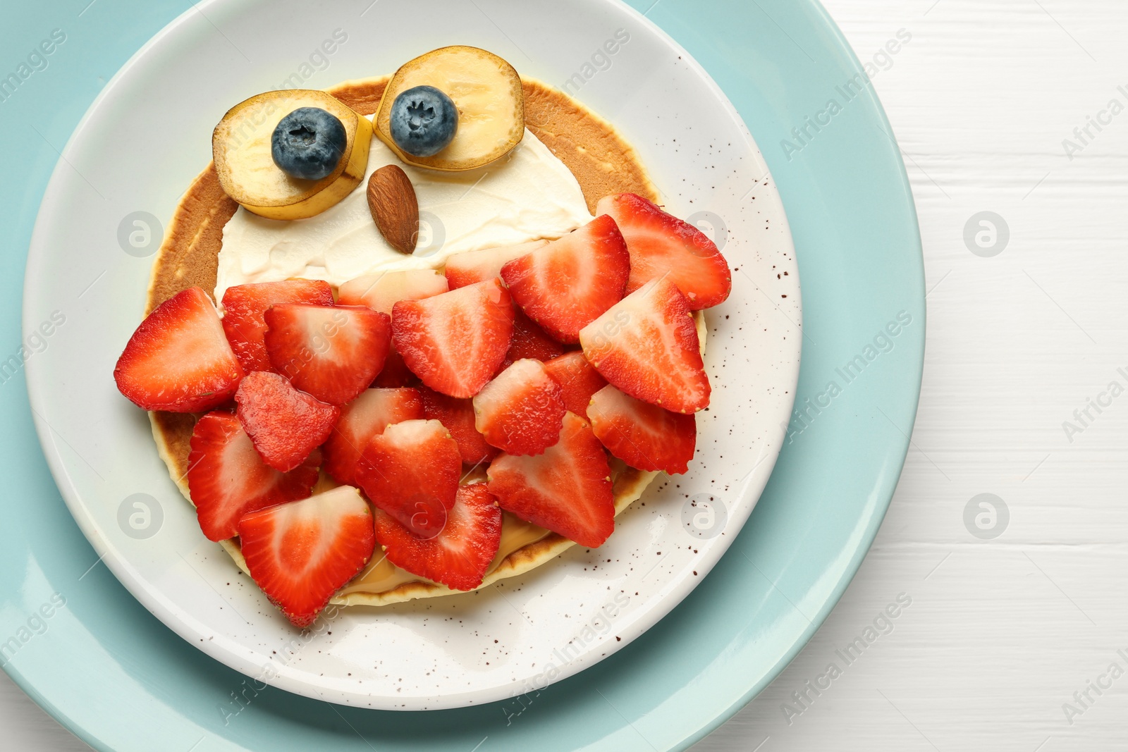 Photo of Creative serving for kids. Plate with cute owl made of pancakes, strawberries, cream, banana and almond on white wooden table, top view