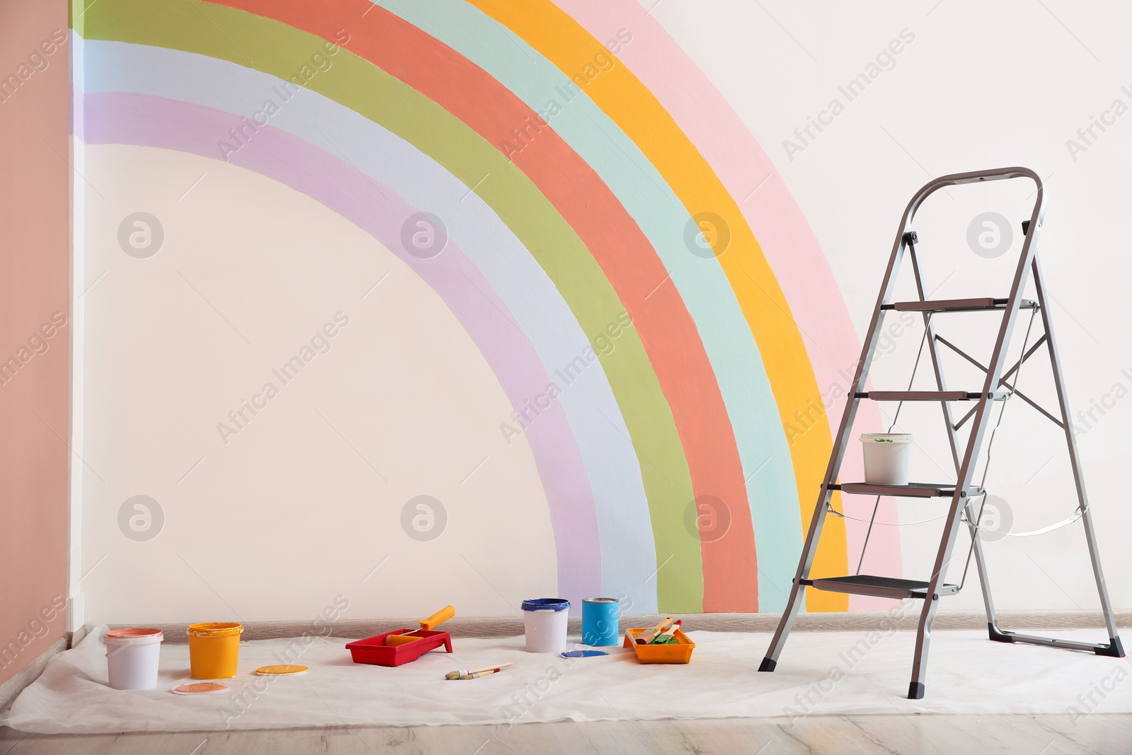 Photo of Different decorator's tools and ladder near wall with painted rainbow indoors