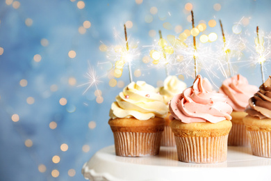 Image of Delicious birthday cupcakes with sparklers on stand against blurred background. Space for text