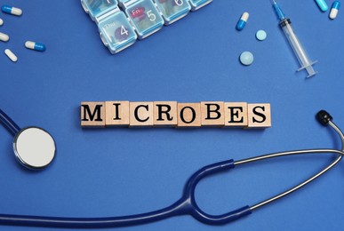 Photo of Word Microbes made with wooden cubes, colorful pills, syringe and stethoscope on blue background, flat lay